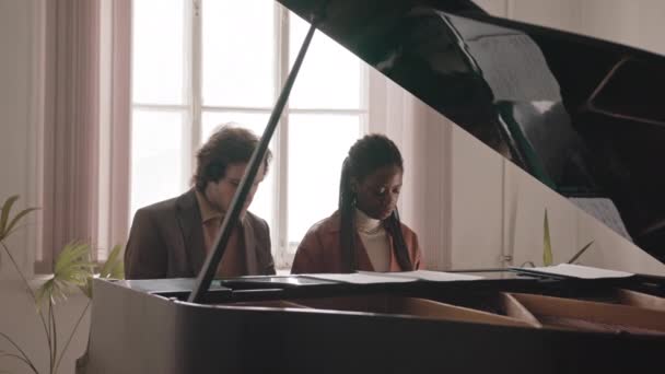 若い白人男性の中低速キーボードの手で楽器で一緒に座っている若いアフリカ系アメリカ人教師と音楽クラスでグランドピアノを演奏することを学ぶ — ストック動画