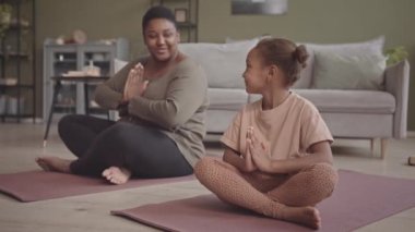 Rahatlamış Afro-Amerikan kıvrımlı kadın ve 5 yaşındaki kızı yoga minderlerinde oturup birlikte meditasyon yapıyorlar.
