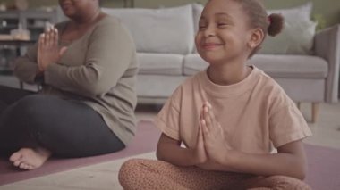 Yavaşça gülümseyen Afro-Amerikalı bir kız yoga minderinde oturmuş dua eden elleri ve gözleri kapalı meditasyon yaparken annesiyle evde.