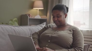 Evdeki kanepede çalışırken dizüstü bilgisayar kullanan Afro-Amerikalı genç bir kadın.