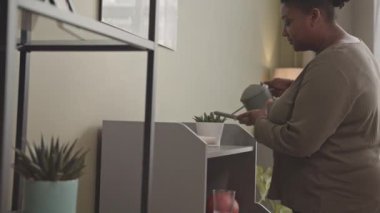 Genç Afro-Amerikan vücudunun orta yavaşlığı. Rahat giyimli kadın. Oturma odasındaki çiçekleri suluyor.