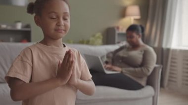 5 yaşındaki Afro-Amerikalı bir kızın oturma odasında meditasyon yapması ve annesinin arka planda çalışırken dizüstü bilgisayar kullanması.