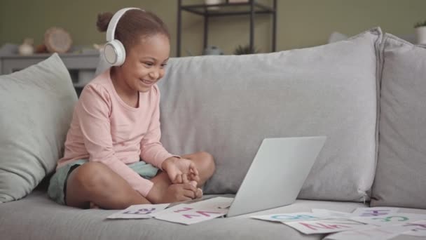 喜びの遅いです5歳のアフリカ系アメリカ人の女の子でヘッドフォンでビデオレッスンを持っていますラップトップ上のソファの上にリビングルームに座って — ストック動画