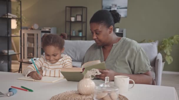 家で勉強するアフリカ系アメリカ人の若い女性と彼女の就学前の娘のウエストアップ遅い コピーブックでアルファベットの手紙を書く練習 — ストック動画