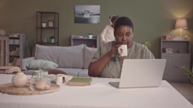 Orta yaşlı, genç, pozitif, Afro-Amerikalı bir kadın laptopla evden çalışırken, kulaklıklı, şakacı küçük kızı arka plandaki koltukta zıplayıp eğleniyor.