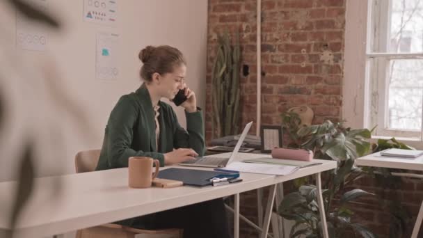 中程度の遅いの若い白人のビジネスマンの女性のスマートフォンに座っている彼女の職場でラップトップの前で彼女の現代的なオフィス — ストック動画