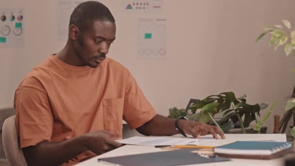 身穿橙色T恤衫的认真的非洲裔美国人坐在办公室里检查财务文件的慢动作 — 图库视频影像
