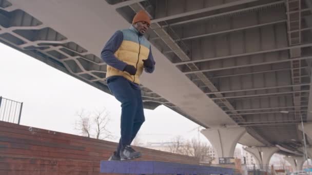 寒い朝に橋の下の屋外ワークアウト中にベンチにジャンプ暖かい活動中のアフリカ系アメリカ人男性の完全な長さの減速 — ストック動画