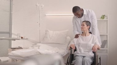 Destekleyici Afrikalı Amerikalı bir doktorun, yetişkin beyaz bir kadını, hastane koğuşunda ameliyat sırasında tekerlekli sandalyeye oturtması.