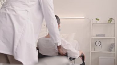 Orta yaşlı Afrikalı Amerikalı doktor, tekerlekli sandalyedeki olgun beyaz kadının hastane yatağında oturup hastalığını veya modern klinikteki ameliyatını atlatmasına yardım ediyor.