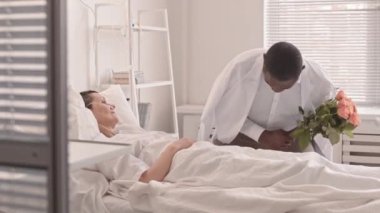 Bir buket çiçekli Afrikalı Amerikalı bir adamın hastanedeki kız arkadaşını ziyaret edişinin orta boy fotoğrafı. Ameliyat sonrası iyileşen bir kadın hastane yatağında yatıyor.