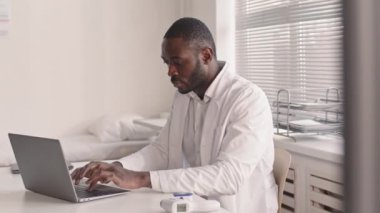 Beyaz laboratuvar önlüklü özgüvenli Afrikalı Amerikalı erkek doktorun modern ofisinde iş yerinde laptopla çalışırken orta derecede yavaşlaması.