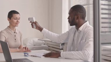 Modern klinikte doktor randevusu almak için gelen kısa saçlı, beyaz bir kadın. Afrikalı Amerikalı uzman hasta sıcaklığını temassız termometreyle ölçüyor.