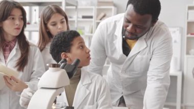 Fen dersinde mikroskop kullanarak laboratuvar önlüklü Afrika kökenli Amerikalı kız öğrencilerin orta yavaşlığı. Bu sırada sınıf arkadaşları ve öğretmenleri yakınlarda durup onları izliyor ve not alıyorlar.