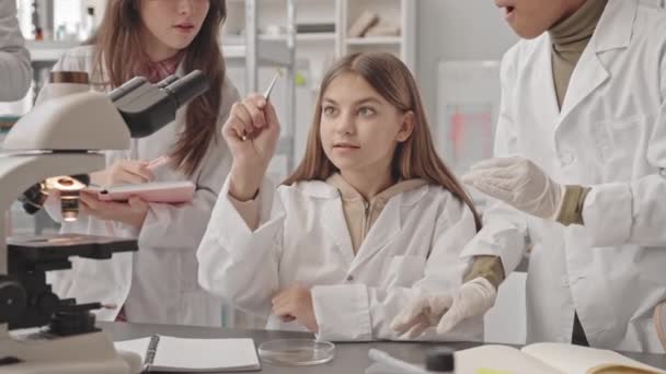 研究室のコートを着た多民族の生徒の中には 科学の授業中に顕微鏡下で見たペトリ皿の認識できないサンプルを調べることができます — ストック動画
