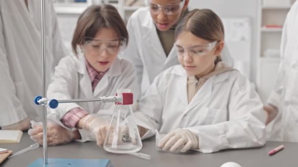 研究室のコートを着た子供たちの中程度のスローモは 液体で茶色の粉をガラスフラスコに入れて化学実験を行い 反応としてフラスコからラボの煙を出す — ストック動画