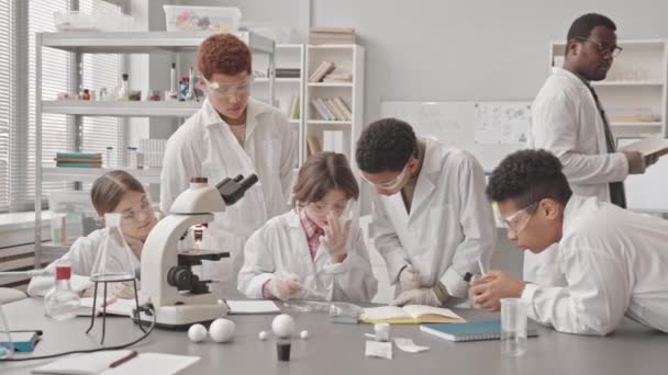 研究室のコートを着たスマートな多民族学生のグループの中低速およびペトリ皿のサンプルを用いた実験を行う保護ガラス現代の教室での科学レッスン — ストック動画