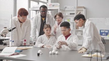 Çok ırklı öğrencilerden oluşan bir grup ve laboratuvar önlüklü Afrikalı Amerikalı öğretmenleri tepkime olarak matarada bolca duman deniyorlar, fen dersinde birlikte eğleniyorlar.