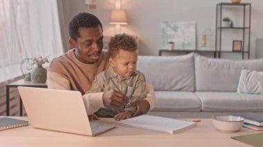 Kilit altındayken laptopta çalışırken küçük oğluna dikkat eden siyahi bir adam.