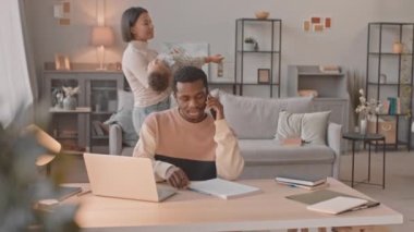 Orta boy Afro-Amerikan iş adamının laptopuyla evden telefon görüşmesi yaparken karısı onu tutarken ve oturma odasında yürürken ağlayan bir çocuğun görüntüsü.