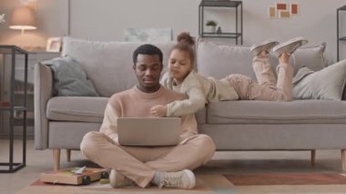 Gündelik giysili Afrikalı bir adam oturma odasında dizüstü bilgisayarda çalışıyor. 8 yaşındaki kızı yakınlarda oynuyor ve tecrit sırasında evde birlikte kalıyorlar.