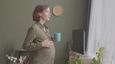 Birbirlerine aşık lezbiyen çiftin yakında ebeveyn olmaya hazırlanmak için evde vakit geçirmesi. Genç beyaz hamile kadın, karısı bebek kıyafetlerini düzenlerken çay içiyor.