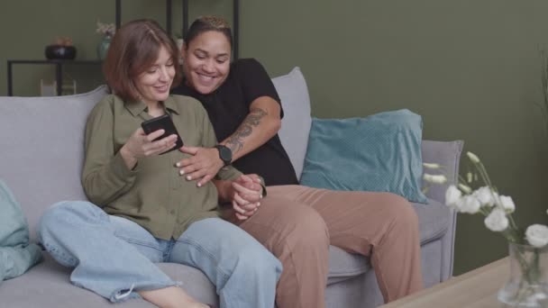 幸せな異人種間のレズビアンカップルの赤ちゃんを待っているのが遅い 2人の女性がソファに座って手をつないで 赤ちゃんの超音波画像をスマートフォンで見て — ストック動画