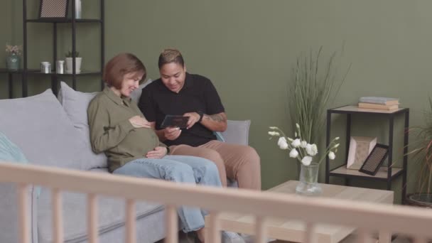 慢吞吞地喜欢跨种族同性恋夫妇看着婴儿超声波图像坐在沙发上的家 分娩时的女人在头顶亲吻她心爱的女性伴侣 — 图库视频影像