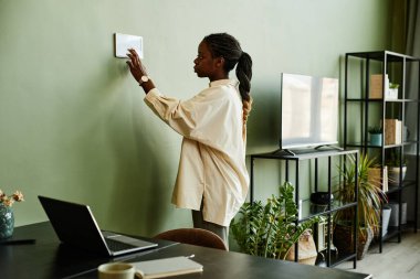 Asgari modern iç mekanda akıllı ev kontrol paneli kullanan Afrikalı Amerikalı genç bir kadının yan görünüm portresi.