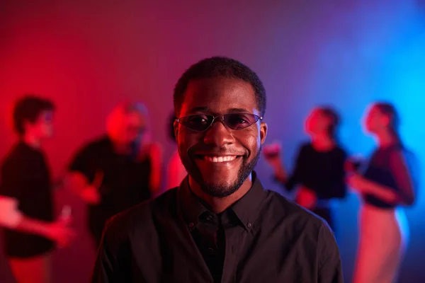 Front view portrait of African American man smiling at camera at party and wearing colored glasses in neon light