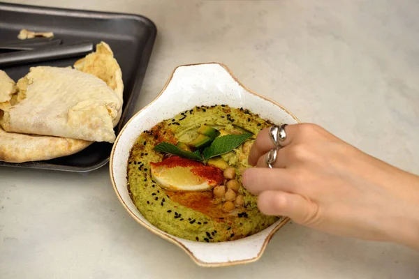 Hummus with avocado in a ceramic pan and Lebanese tortilla. Traditional Jewish cuisine. A woman\'s hand dips piece of bread into hummus. Close-up