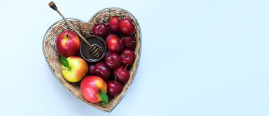 Fruit for the Jewish New Year. Red apples, plums and honey in straw basket made in the shape of heart on green background. Copy space
