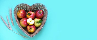 Red apples and ears of wheat in straw basket made in the shape of heart on green background. Religious holidays concept - Jewish New Year or Transfiguration of the Lord. Copy space