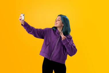 Cheerful nice young girl with blue hair taking selfie on cellphone and smiling. Isolated on yellow baclground