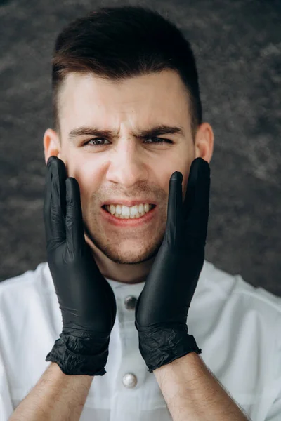 A male doctor in black gloves puts on holds on to his face and suffers from pain