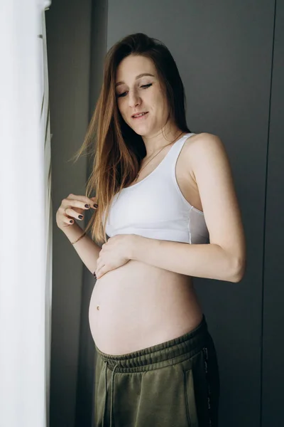 Έγκυος Μακρυμάλλης Κορίτσι Πράσινο Παντελόνι Στέκεται Κοντά Στο Παράθυρο — Φωτογραφία Αρχείου