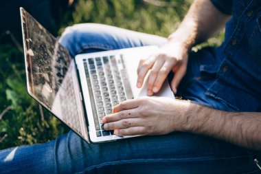 Yaz yaşam tarzı yakışıklı sakallı Avrupalı iş adamı mavi kot ceketli yeşil çimlerde oturuyor elinde Macbook ve daktilo ile