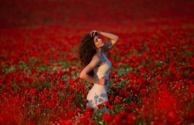 Güzel, ince, uzun saçlı, beyaz elbiseli esmer kadın kırmızı bir çiçek tarlasının ortasında duruyor.. 
