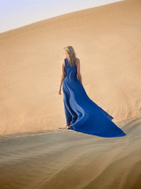 Beautiful long haired blonde woman in long blue flying dress walks away among dunes in the middle of a desert.