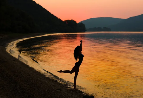 日落时 完美性感的女人在河边跳舞和摆姿势 橙色背景 黑色身体轮廓 图库照片