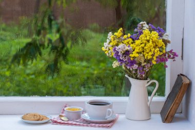 Eski bir kitap, renkli bir buket çiçek ve arka planda beyaz bir fincan çay. Yaz günü bahçede yağmur damlaları var. Kapatın, boşluğu kopyalayın.