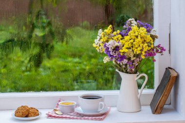 Eski bir kitap, renkli bir buket çiçek ve arka planda beyaz bir fincan çay. Yaz günü bahçede yağmur damlaları var. Kapatın, boşluğu kopyalayın.