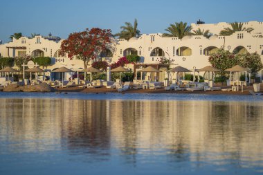 Binalar, güneş panelleri, şemsiyeler ve palmiye ağaçları, Mısır, Afrika 'daki Sharm El Sheikh tatil beldesinde sabahleyin sahildeki sakin Kızıl deniz suyuna yansıyor. Karantina sırasında boş plajlar