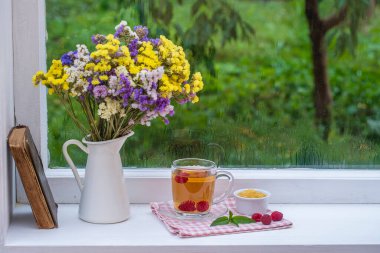 Eski bir kitap, renkli bir buket çiçek ve arka planda beyaz bir fincan çay. Yaz günü bahçede yağmur damlaları var. Kapatın, boşluğu kopyalayın.
