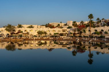Binalar, güneş panelleri, şemsiyeler ve palmiye ağaçları, Mısır, Afrika 'daki Sharm El Sheikh tatil beldesinde sabahleyin sahildeki sakin Kızıl deniz suyuna yansıyor. Karantina sırasında boş plajlar