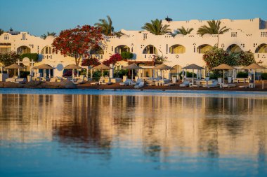 Binalar, güneş panelleri, şemsiyeler ve palmiye ağaçları, Mısır, Afrika 'daki Sharm El Sheikh tatil beldesinde sabahleyin sahildeki sakin Kızıl deniz suyuna yansıyor. Karantina sırasında boş plajlar