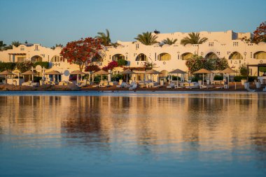 Binalar, güneş panelleri, şemsiyeler ve palmiye ağaçları, Mısır, Afrika 'daki Sharm El Sheikh tatil beldesinde sabahleyin sahildeki sakin Kızıl deniz suyuna yansıyor. Karantina sırasında boş plajlar