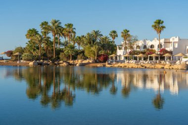 Binalar, güneş panelleri, şemsiyeler ve palmiye ağaçları, Mısır, Afrika 'daki Sharm El Sheikh tatil beldesinde sabahleyin sahildeki sakin Kızıl deniz suyuna yansıyor. Karantina sırasında boş plajlar