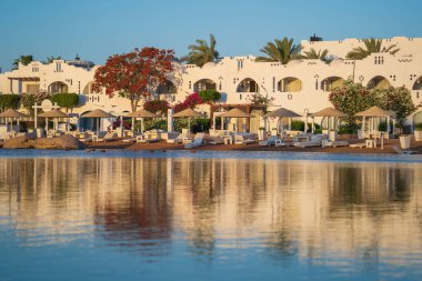 Gündoğumunda, Mısır 'ın Sharm El Sheikh tatil beldesindeki sakin deniz suyuna binalar, güneşlikler ve şemsiyeler yansıtılır.
