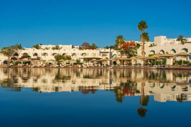 Gündoğumunda, Mısır 'ın Sharm El Sheikh tatil beldesindeki sakin deniz suyuna binalar, güneşlikler ve şemsiyeler yansıtılır.